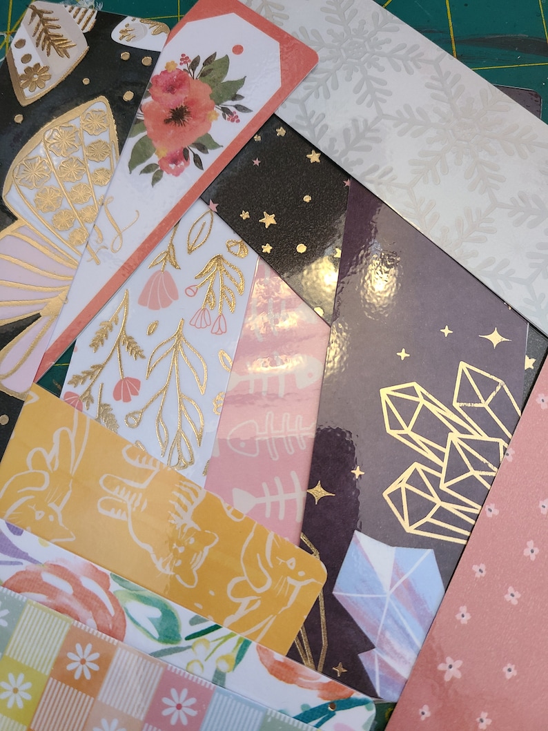 May include: A collection of colorful bookmarks with various designs, including floral patterns, geometric shapes, and animal illustrations. The bookmarks are made of paper and have a glossy finish.