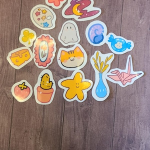 Op de afbeelding: Een verzameling van 14 kleurrijke stickers met verschillende schattige illustraties, waaronder een bloem, een geest, een kat, een cactus, een ster, een vaas met tarwe, een papieren kraanvogel, een stuk kaas, een palet, een werveling, een snoepje, een klavertjevier, een fotolijst en een aardbei.