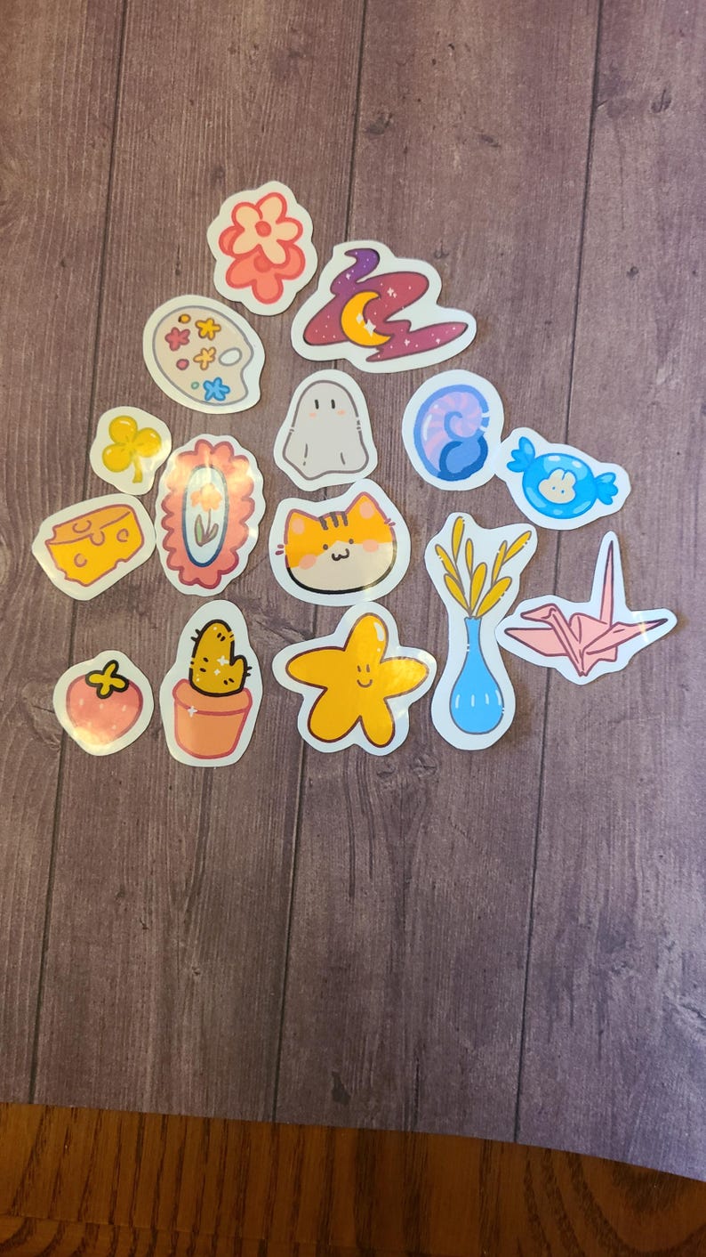Op de afbeelding: Een verzameling van 14 kleurrijke stickers met verschillende schattige illustraties, waaronder een bloem, een geest, een kat, een cactus, een ster, een vaas met tarwe, een papieren kraanvogel, een stuk kaas, een palet, een werveling, een snoepje, een klavertjevier, een fotolijst en een aardbei.