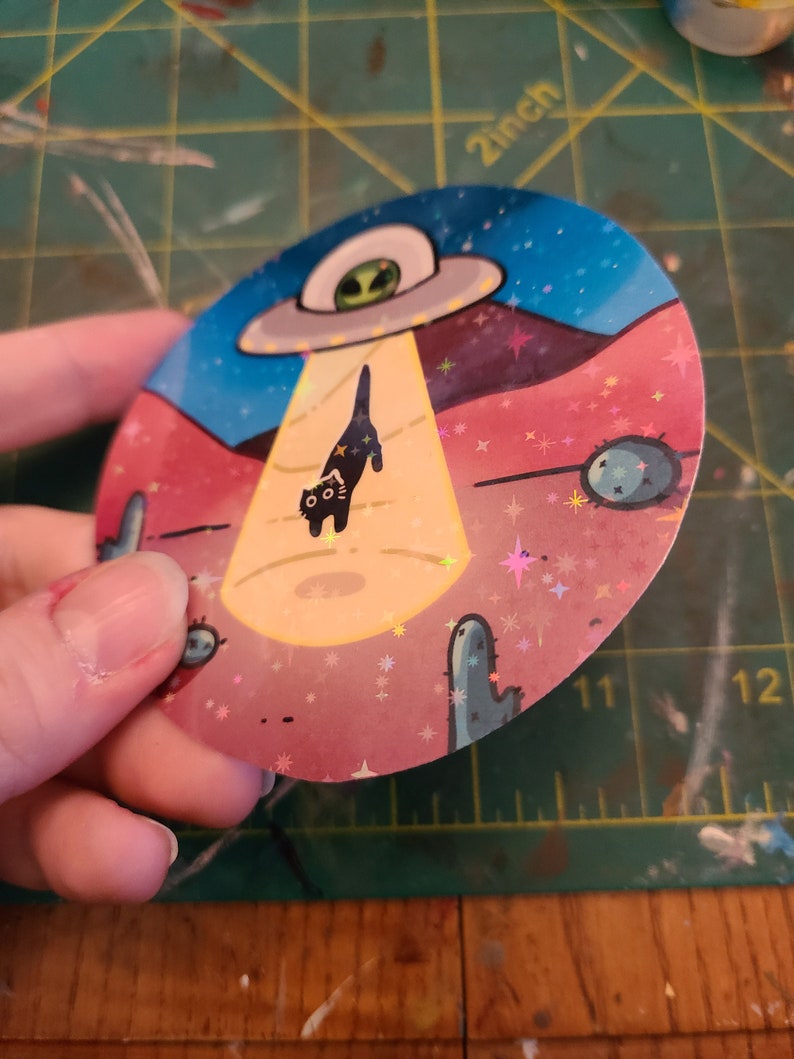 May include: A round sticker with a black cat being abducted by a UFO. The sticker has a pink, blue, and yellow background with glitter and stars. The cat has a surprised expression on its face.