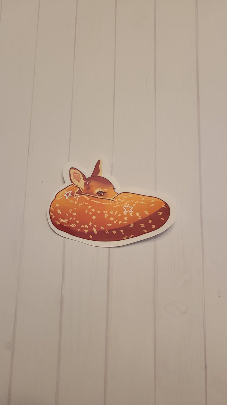 May include: A sticker featuring a curled-up fawn with a speckled orange and brown coat. The fawn's head is tucked in, and it has a white border. The sticker is on a white background with vertical wood grain.