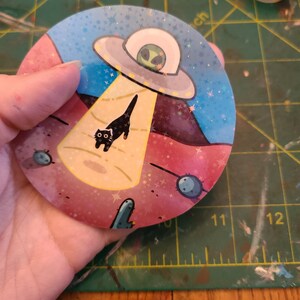 May include: A round sticker with a pink, blue, and yellow background. A black cat is walking towards a flying saucer with a green alien inside. The sticker has a glittery finish and stars scattered across the background.