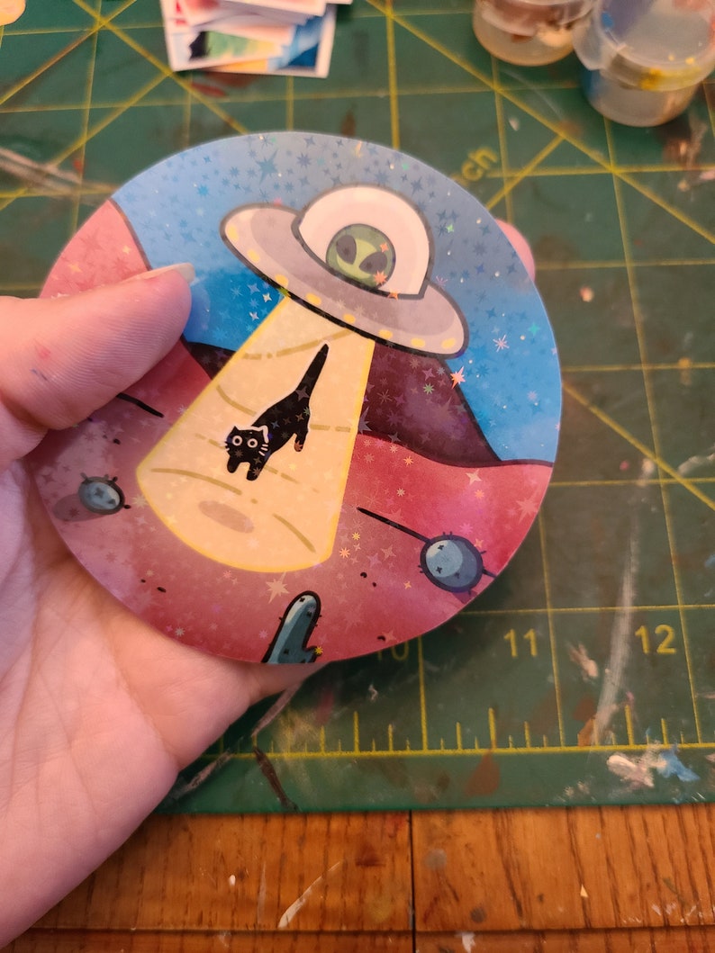 May include: A round sticker with a pink, blue, and yellow background. A black cat is walking towards a flying saucer with a green alien inside. The sticker has a glittery finish and stars scattered across the background.