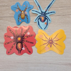May include: Four colourful spider-shaped stickers. One is blue with a black spider, another is red with a brown spider, and the other two are yellow and blue with spiders. Each sticker has star accents.