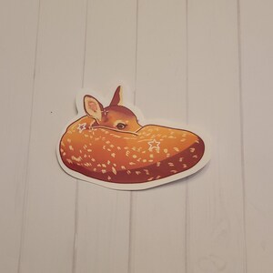 May include: A sticker featuring a curled-up fawn with a speckled orange and brown coat. The fawn's head is tucked in, and it has a white border. The sticker is on a white background with vertical wood grain.