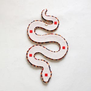 May include: A white felt snake-shaped wall hanging with a textured, multi-colored border. The snake has a gold ring attached and four red square accents. The design is a whimsical, decorative piece.