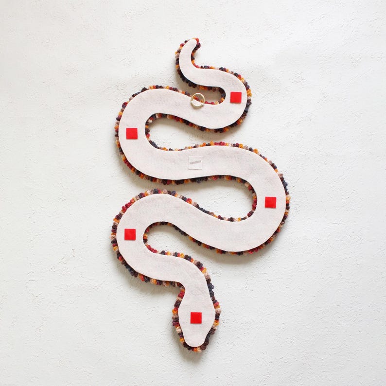 May include: A white felt snake-shaped wall hanging with a textured, multi-colored border. The snake has a gold ring attached and four red square accents. The design is a whimsical, decorative piece.