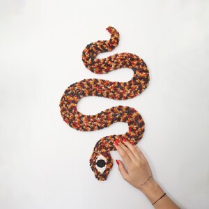 May include: A handmade, plush snake in shades of brown, orange, and black, coiled on a white surface. The snake's head features a large, black and white eye. The snake is made of a soft, textured material.