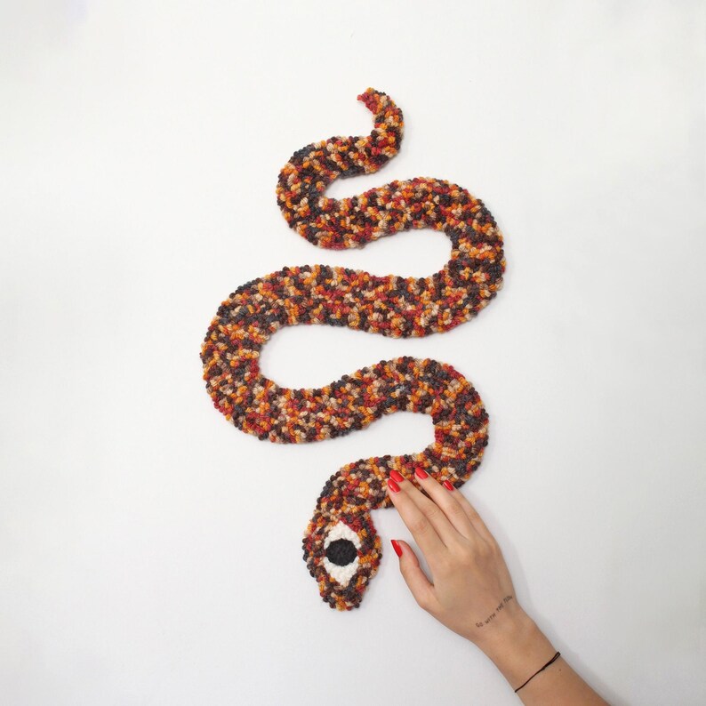 May include: A handmade, plush snake in shades of brown, orange, and black, coiled on a white surface. The snake's head features a large, black and white eye. The snake is made of a soft, textured material.