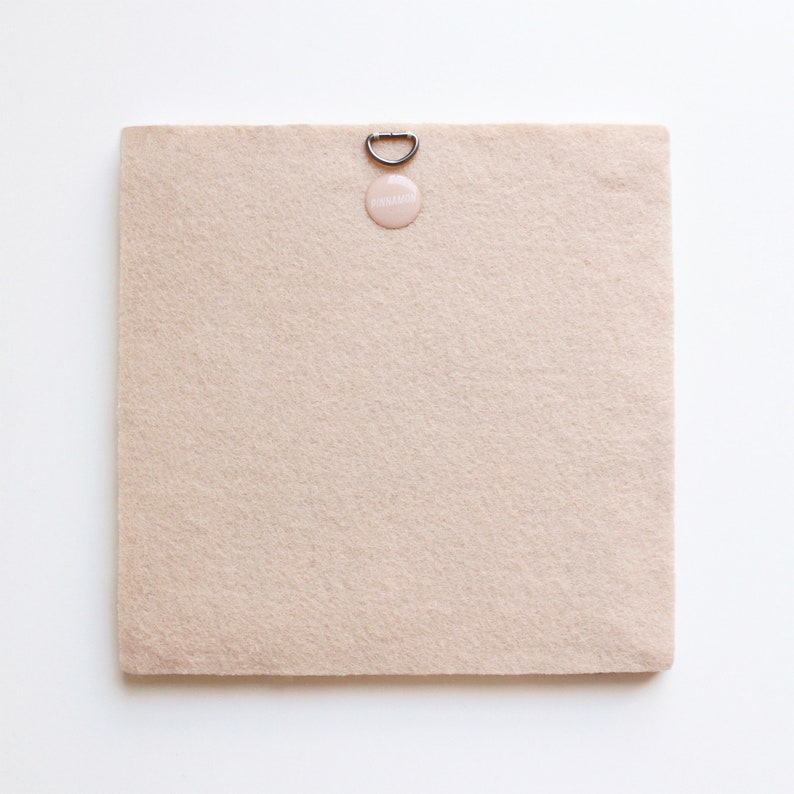 May include: A beige felt board with a silver hanging hook and a pink round sticker that says "remarkable".