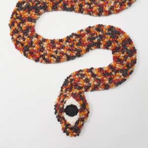 May include: A handmade, knitted snake in shades of brown, orange, and red. The snake's head features a black and white eye, and the body is curved in a playful, winding shape. The textile art piece is set against a white background.