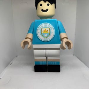 May include: A large, light blue figurine of a soccer player in a Manchester City uniform. The figure has a black hair, a smiling face, and the Manchester City logo on the chest. The figure is wearing white shorts and black shoes.