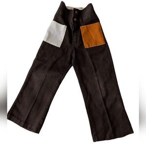May include: Dark brown pants with high waist and two buttons. The pants have two square patches, one white and one orange. The pants are made of a corduroy-like material.