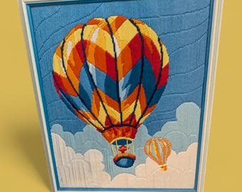 May include: A framed needlepoint artwork featuring two hot air balloons against a blue sky with white clouds. The larger balloon is a vibrant mix of orange, yellow, red, and blue. The frame is white.