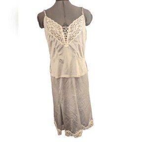 May include: A vintage-style slip dress in a soft, silvery-gray hue. The bodice features a delicate ivory lace trim at the neckline and a matching lace detail at the hem. The dress is displayed on a mannequin.