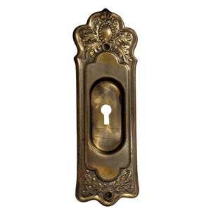 May include: Ornate brass door plate with a keyhole. The rectangular plate features decorative floral and scrollwork designs at the top and bottom. The keyhole is oval-shaped. The brass has a warm, aged patina.
