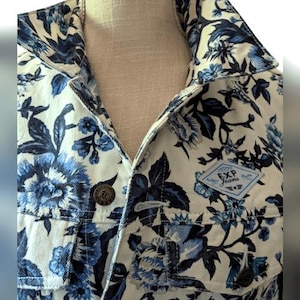 May include: A denim jacket with a white background and a blue floral pattern. The jacket has a button-down front, two chest pockets, and a small EXP Jeans patch. The jacket is on a mannequin.