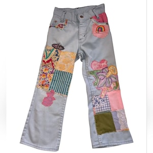 May include: Light blue denim jeans with colorful patchwork and embroidered floral and butterfly designs. The jeans feature a straight leg cut and a button and zipper closure. Patches include floral, striped, and gingham patterns.