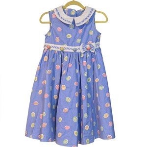 May include: A sleeveless blue dress with a white collar and a bow at the waist. The dress is decorated with colorful, circular patterns and small buttons. The dress is hanging on a gold hanger.
