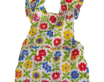 May include: White romper with a colorful floral pattern featuring red, yellow, and blue flowers. The romper has ruffled sleeves and white buttons. The overall design is a cheerful, vintage-inspired style.