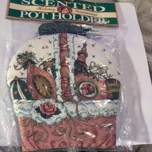May include: A scented holiday pot holder with a festive design. The pot holder features a pink and white floral pattern with images of ornaments and a decorative basket. The top of the pot holder has a green and white polka dot bow. The packaging reads "Scented Holiday Trimmings".