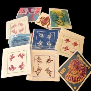 May include: A collection of square paper prints with various designs. Some prints feature red frogs, blue and white spiral patterns, and a corn graphic. Other prints display ancient Egyptian imagery and other abstract designs.