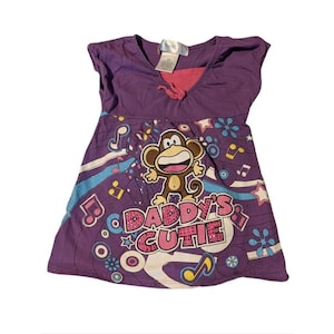 May include: A purple short-sleeved dress featuring a graphic of a brown monkey and the text DADDY'S CUTIE in pink glittery letters. The design includes colorful musical notes, stars, and circular patterns against a purple background.