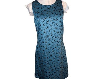 May include: A sleeveless, knee-length dress in a teal color, adorned with a delicate pattern of small black floral designs. The dress has a round neckline and a fitted silhouette, displayed on a mannequin.