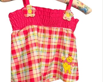 May include: A pink and plaid tank top with flower accents and Winnie the Pooh graphic. The top has a smocked bodice and flower details on the straps. The bottom half features a red, white, yellow, and green plaid pattern.