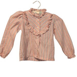 May include: A vintage button-down shirt with a plaid pattern in peach, white, and blue. The long-sleeved shirt features a ruffled collar and cuffs, and is hanging on a white hanger. The shirt has a classic, feminine design.
