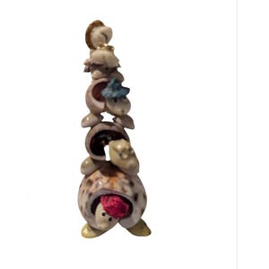 May include: A whimsical stack of seashell figurines, each resembling a turtle. The turtles are in various shades of beige, brown, and green, with a pop of red and blue. The stacked arrangement creates a unique decorative piece.