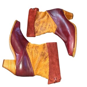 May include: A pair of burgundy leather boots with a pointed toe and wooden heels. The boots feature a tan and orange suede panel on the upper shaft and a dark red suede cuff. The heels are approximately 3 inches high.