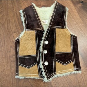 May include: A vintage brown and tan suede vest with a cream-colored shearling lining. The vest features a three-button closure and white stitching details. The vest has a retro style and is likely for a child.