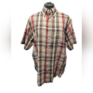 May include: A short-sleeved button-down shirt in a plaid pattern. The shirt features a button-down collar and a chest pocket. The plaid design incorporates red, beige, navy, and olive green colors. The shirt is displayed on a mannequin.
