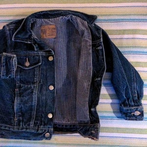 May include: Dark blue denim jacket with silver buttons and a striped interior lining. The jacket has two chest pockets and buttoned cuffs. A brown label is sewn inside the jacket.