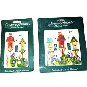 May include: Two decorative ceramic light switch covers featuring hand-painted birdhouse designs. Each cover has a white background with colorful birdhouses, flowers, and the text "Creative Accents Brush Strokes" and "Individually Hand-Painted".