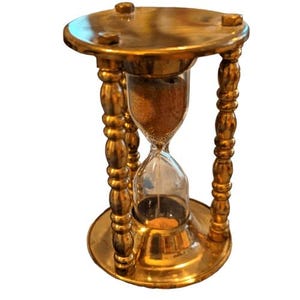 May include: A vintage hourglass with a brass frame and a clear glass center. The hourglass is supported by four ornate legs and has a circular base and top. The sand inside is a light brown color. A decorative item.
