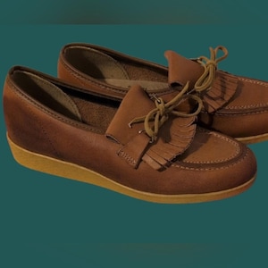 May include: A pair of brown leather loafers with a wedge heel. The shoes have a fringed detail on the vamp and tan-colored soles. The laces are tied in a bow.