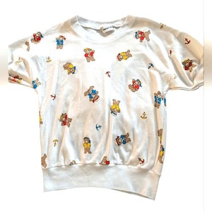 May include: White short-sleeved sweatshirt with a pattern of cartoon bears in sailor outfits and small blue anchors. The bears are in various colors, including red, blue, and yellow. The sweatshirt has a crew neck and a ribbed hem and cuffs.