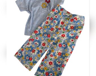 May include: A light blue short-sleeved shirt and matching pants set. The shirt has a small floral design. The pants feature a blue and white checkered pattern with red, yellow, and white flowers. The outfit is likely for a child.