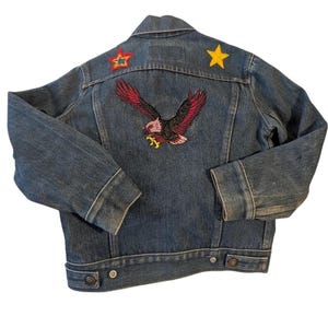 May include: A blue denim jacket with an embroidered eagle design on the back. The jacket features red and yellow star patches on the shoulders. The eagle has red and black wings and yellow talons.