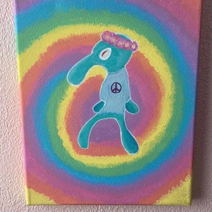 Peut inclure: Une toile peinte à la main représentant un personnage de dessin animé vert avec un signe de paix sur un t-shirt bleu et une couronne de fleurs. Le personnage se tient devant un fond tie-dye coloré.