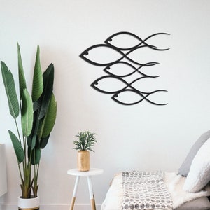 May include: Black metal wall art featuring a school of fish design. The fish are stylized and abstract, with flowing lines and curves. The art is mounted on a white wall, with a green plant and a white side table in the foreground.
