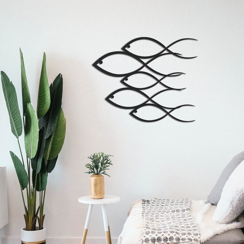 May include: Black metal wall art featuring a school of fish design. The fish are stylized and abstract, with flowing lines and curves. The art is mounted on a white wall, with a green plant and a white side table in the foreground.