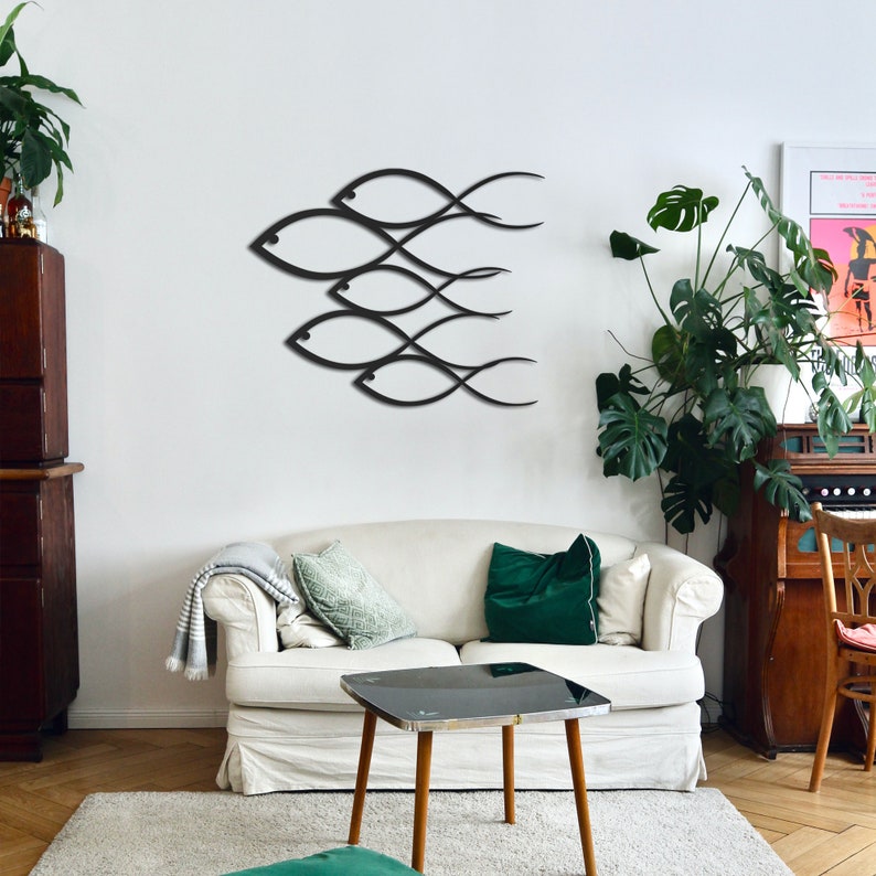 May include: A white couch with green throw pillows in a living room with a black metal wall sculpture of fish. The sculpture is on a white wall. There is a small table in front of the couch and a potted plant in the corner.