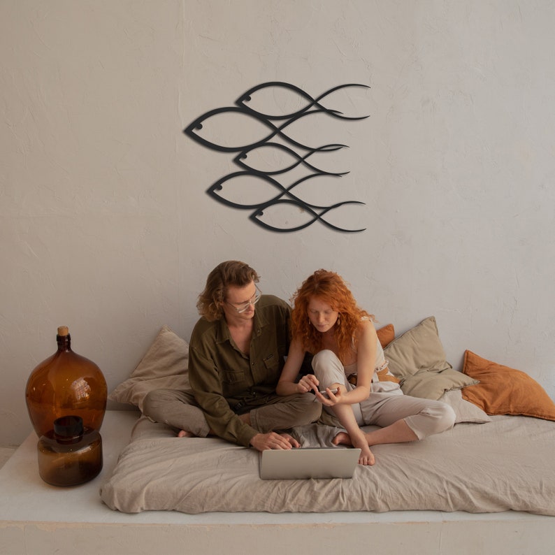 May include: A couple sits on a bed with beige linens and pillows, looking at a laptop computer. A black metal wall sculpture of fish is on the wall behind them.