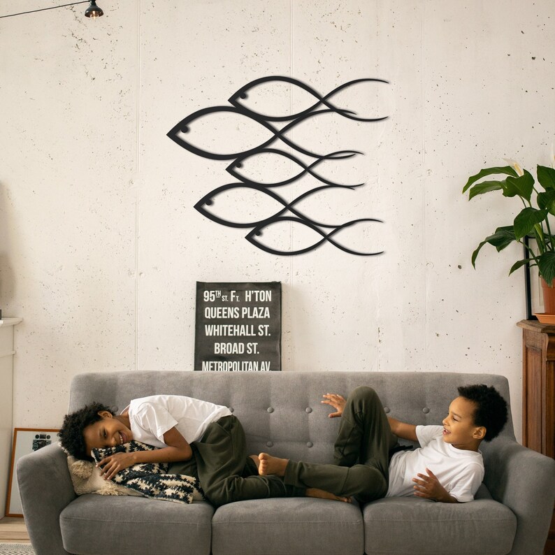 May include: Black metal wall art depicting a school of fish in a minimalist style. The art is mounted on a white wall above a gray couch. Two children are sitting on the couch.