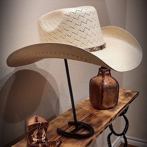 May include: A white straw cowboy hat with a brown leather band is displayed on a metal stand. The hat is sitting on a wooden shelf with a brown leather bottle and a small cowboy boot.