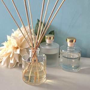 May include: Clear glass diffuser bottles with wooden reeds. One bottle is filled with a light yellow liquid, and two other bottles are visible. A white flower and green leaves are in the background. The bottles have gold-colored caps.