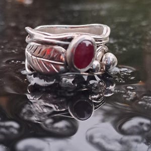 Könnte beinhalten: Ein silberner Ring mit einem roten Edelstein und Blattdetails. Der Ring liegt auf einer feuchten Oberfläche.